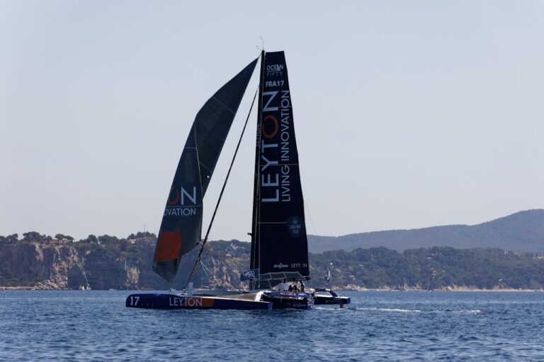 A sleek catamaran with tall sails races on open sea waters, set against a rocky coastal backdrop.